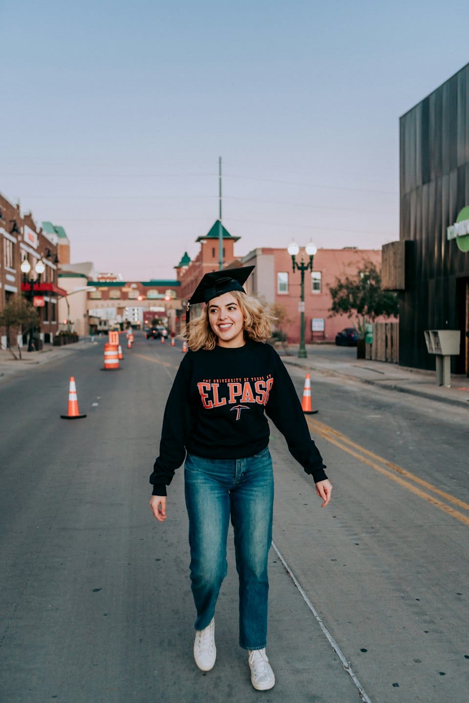 A smiling graduate walks down a city street at dusk.