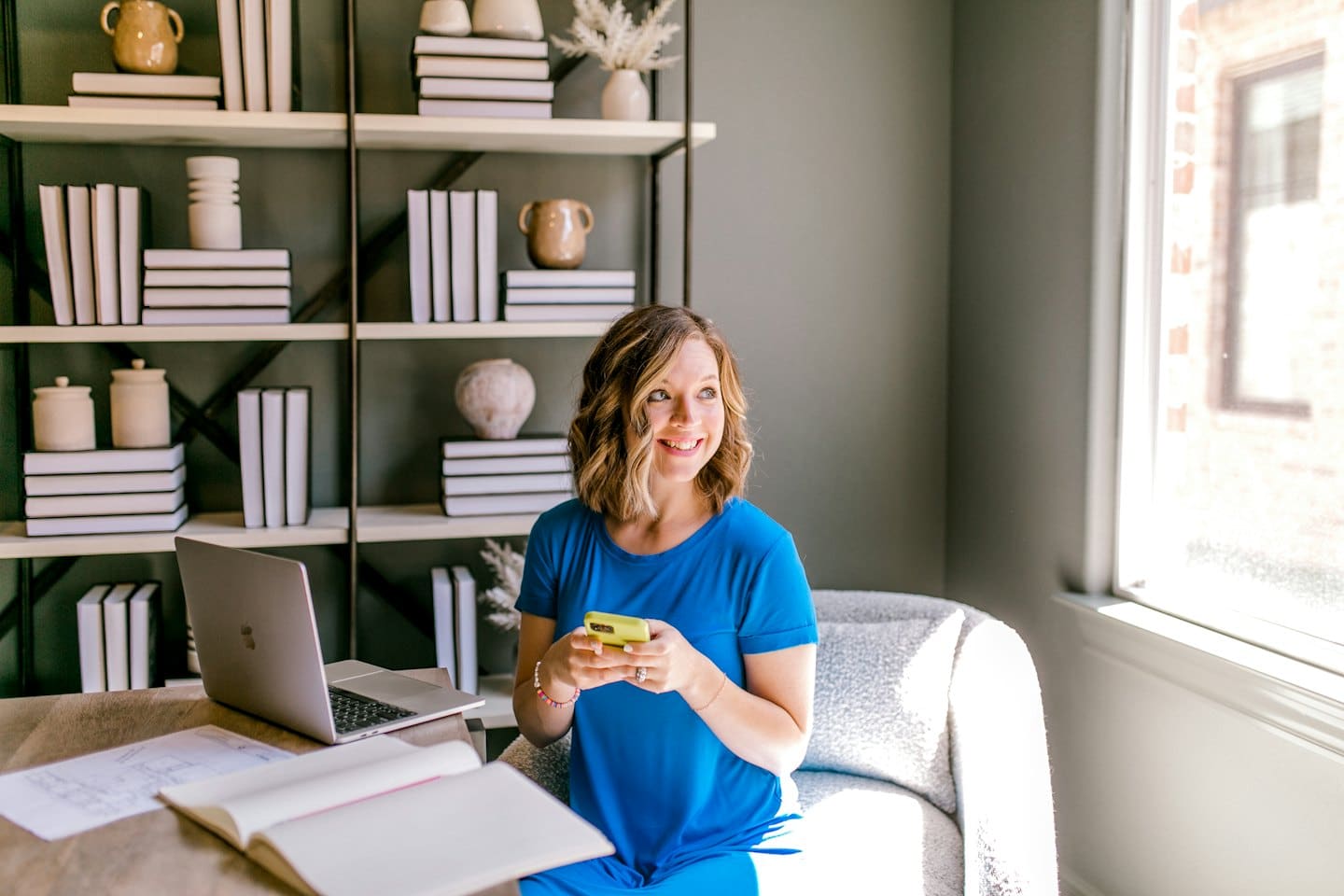 Woman in blue dress uses smartphone at desk with laptop and bookshelf.